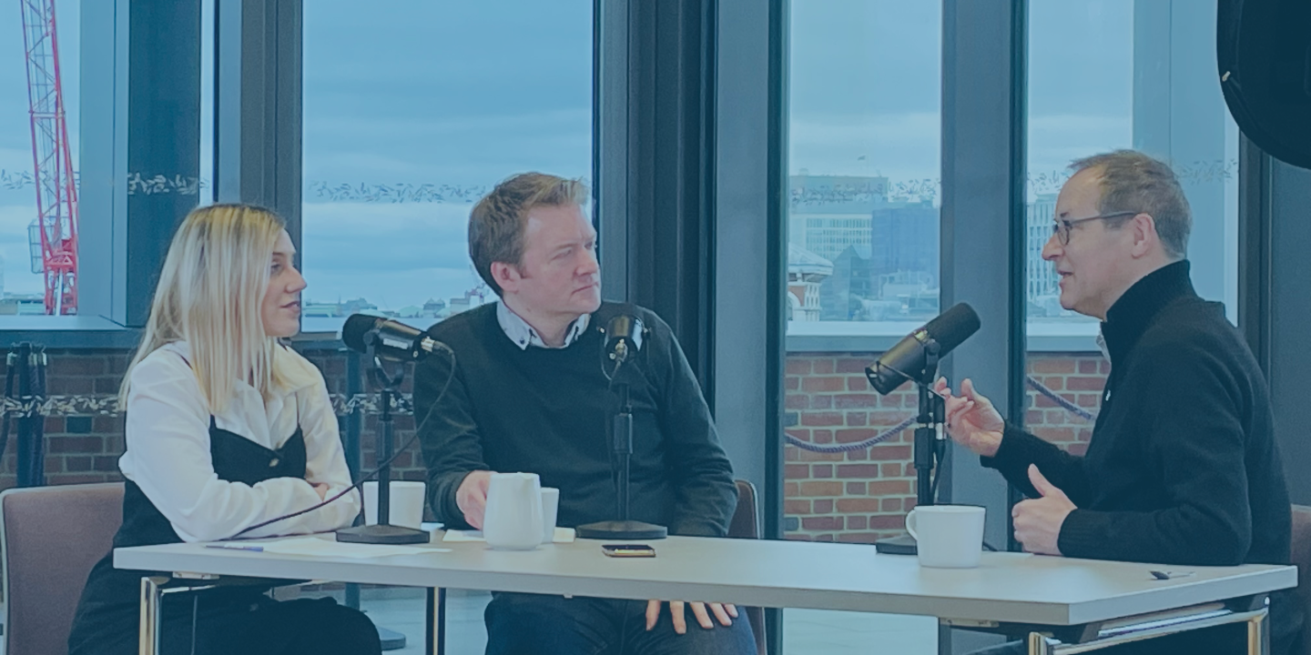 Three people sit around a table recording in front of microphones, one gestures with their hand