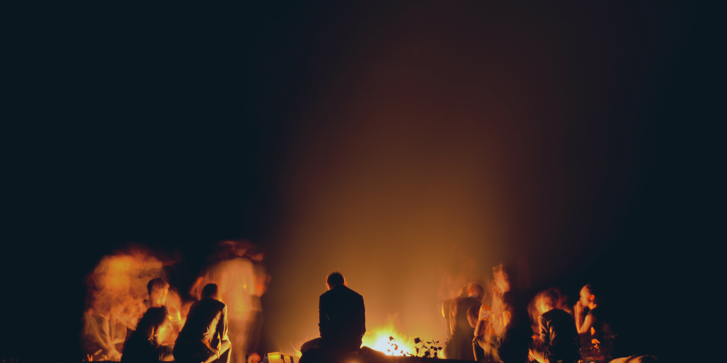 a group of people crowd round a campfire backlighting them in silhouette.