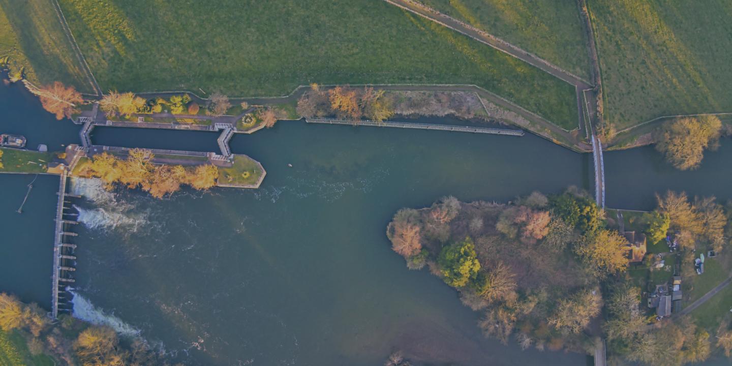 A bird's eye view of a river, with a lock and weir to the left and an island with two bridges to the right.