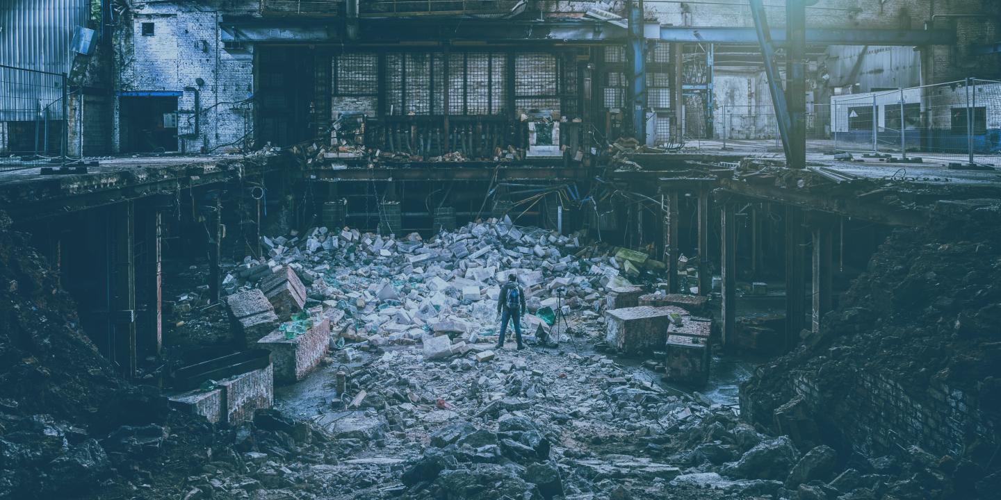 A photographer, standing next to a tripod, atop a pile of rubble is a destroyed factory.