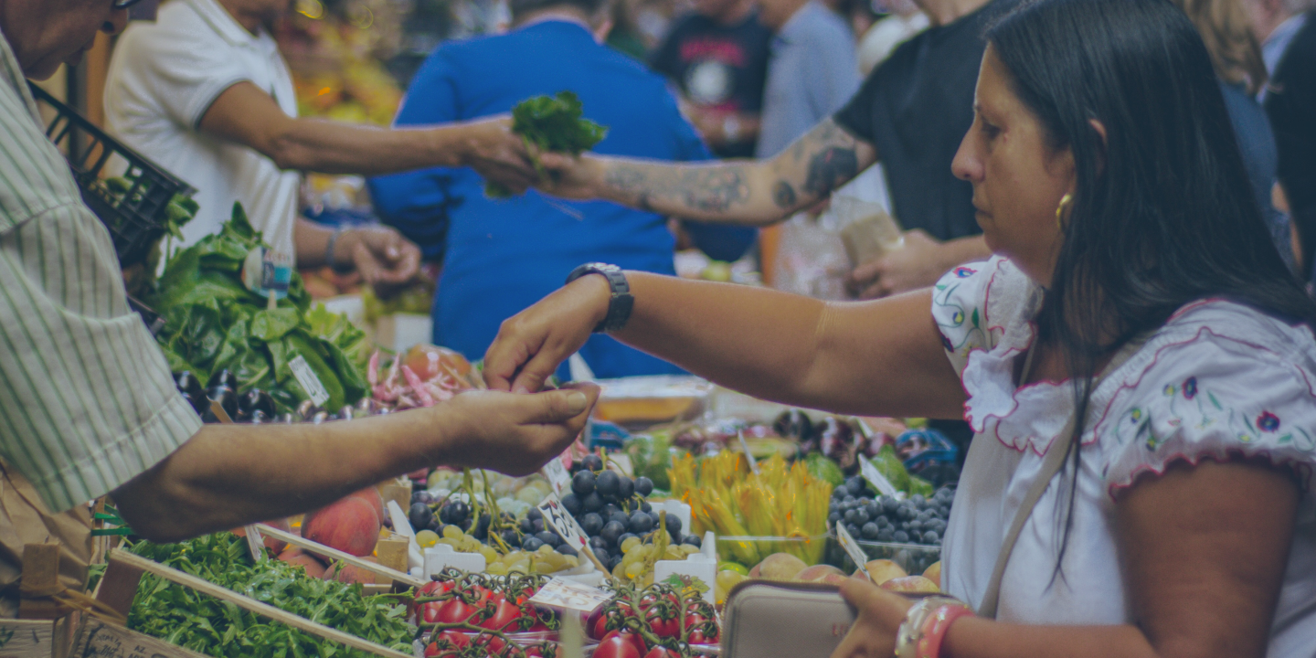 Marketer stall traders and customers reach out arms and hands to exchange fruit and money above piles of produce.