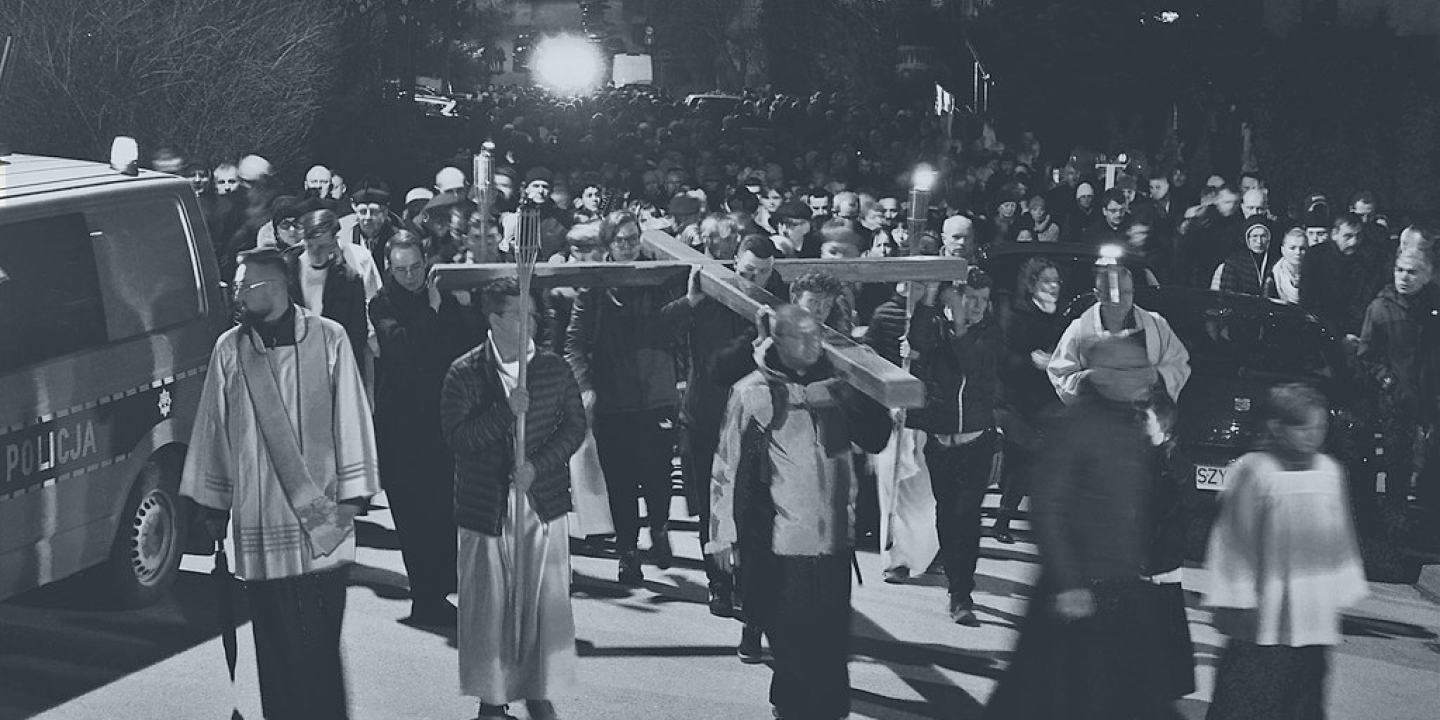 A Good Friday procession of people and priests hold a cross horizontal above their heads.