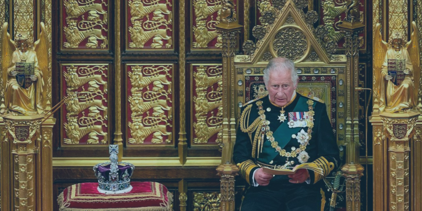 A uniformed Prince Charles sits on a throne reading a speech, beside a crown resting on a cushion