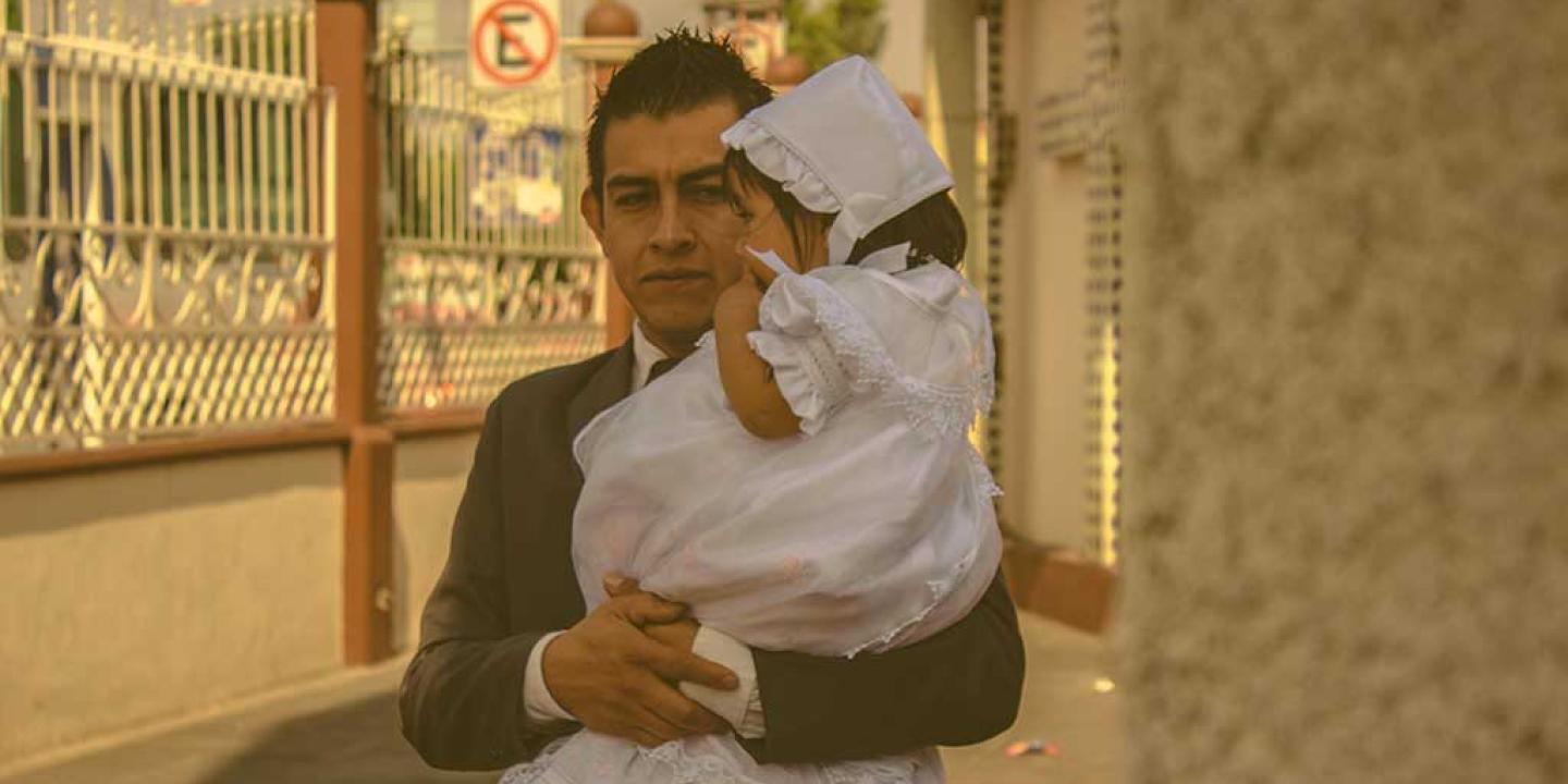 A father wearing a suit carries his child who is dressed for a christening in white.