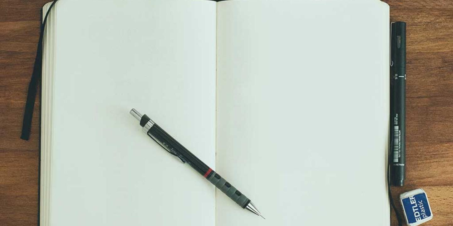 A notebook is open at two blank pages. a pen rests across the page.s.