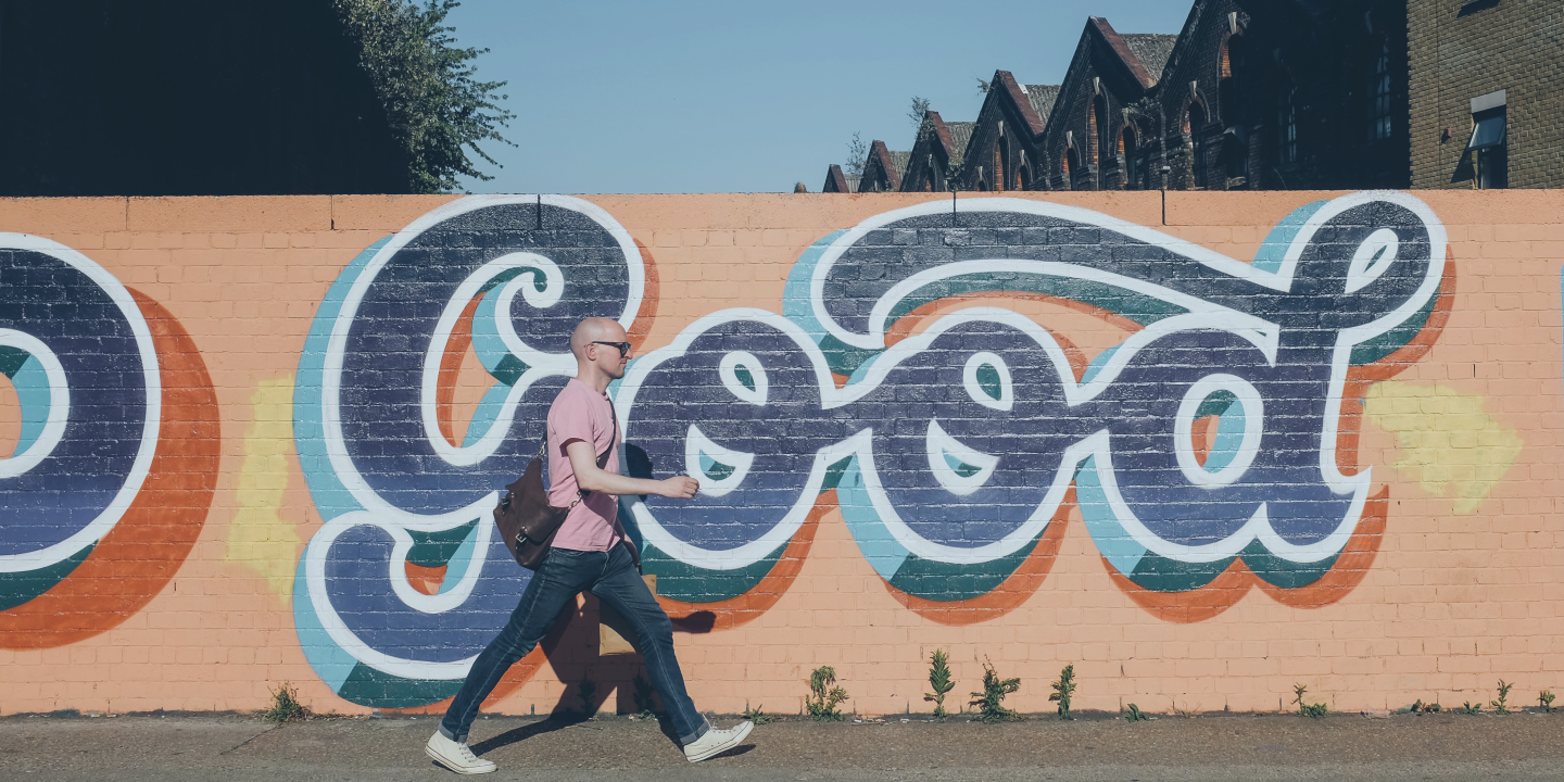 A man walks along a street past a orange wall with a huge 'Good' written in cursive script on it.