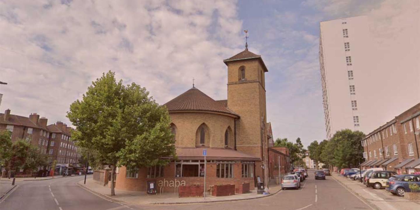 A neo-Romanesque church sits at the acute corner of two roads. To its side a tower block rises over a row of low-rise flats.