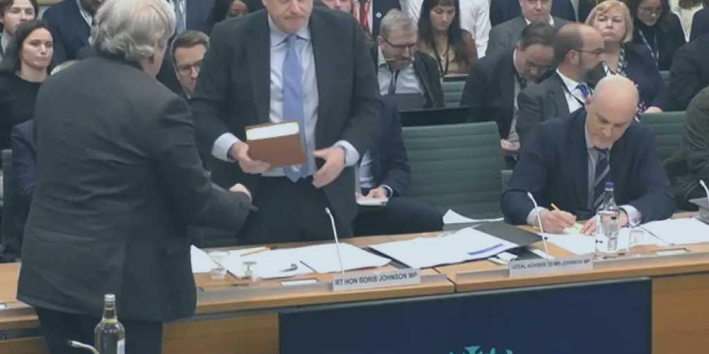 A politican stands holding a bible, in front of a committee room table. Behind him an audience waits expectantly