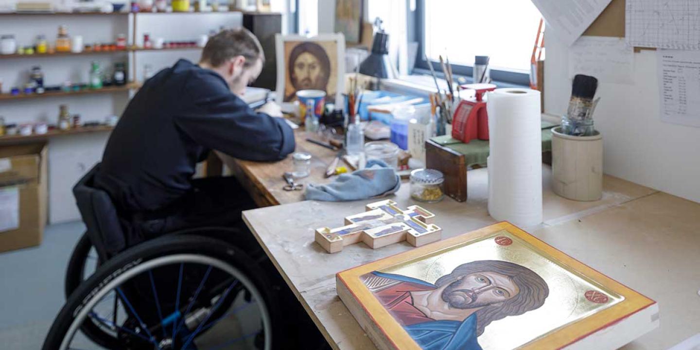 A monk in a wheekchair works on an icon in an art studio. In the foreground is a completed icon. 