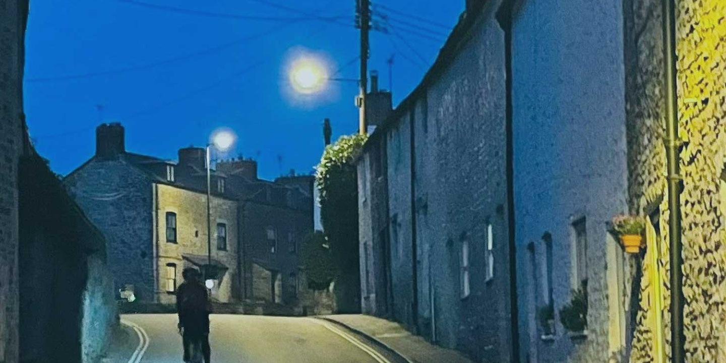 A cyclist ascends a village street between stone-built houses as twilight turns to night