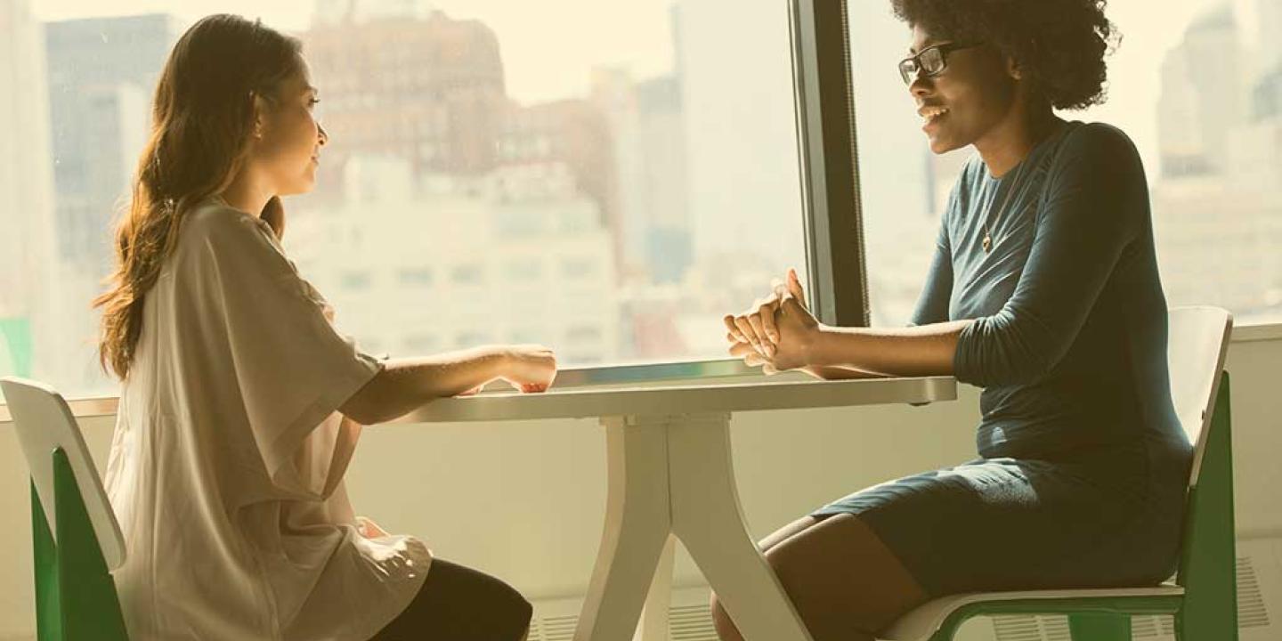 Two people sit at a table with their hands resting on top of it. One speaks to the other