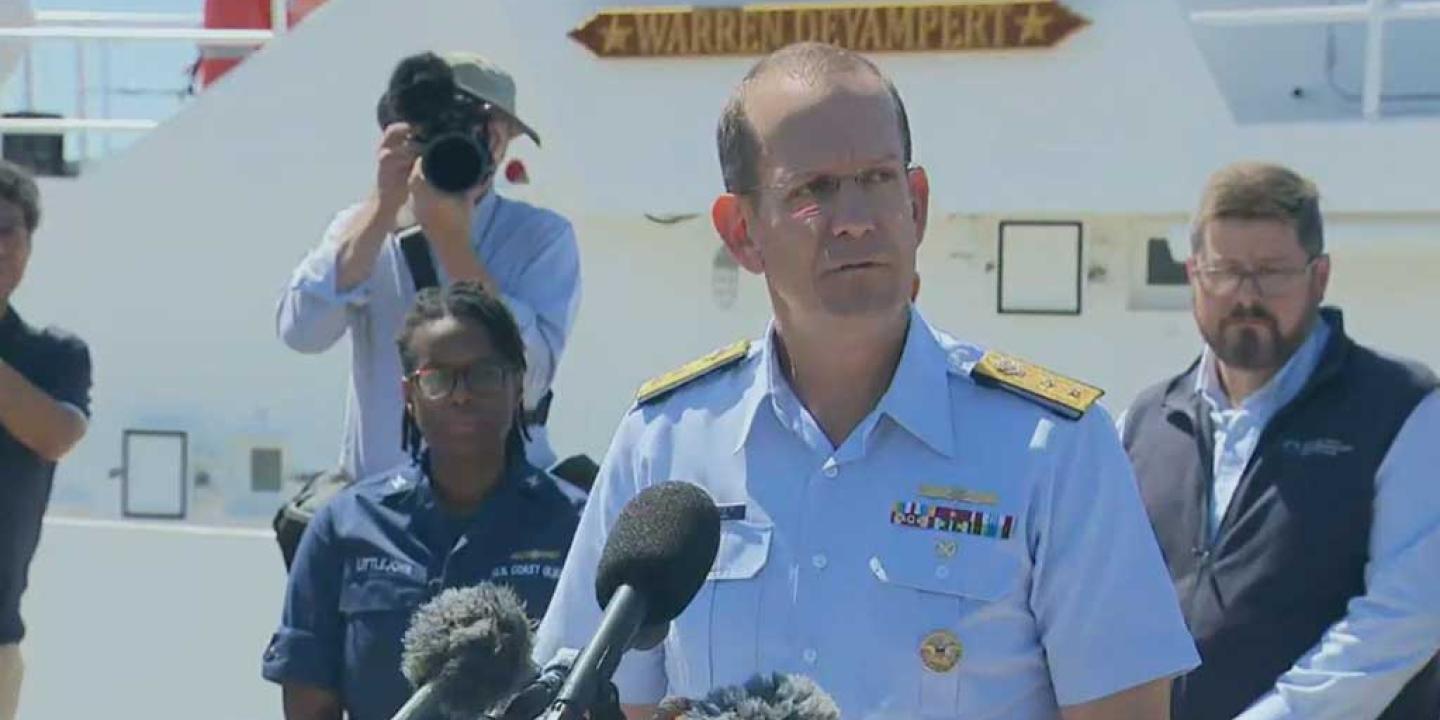 A Coast Guard officer gives a press conference while looking grim-faced. Others look on.