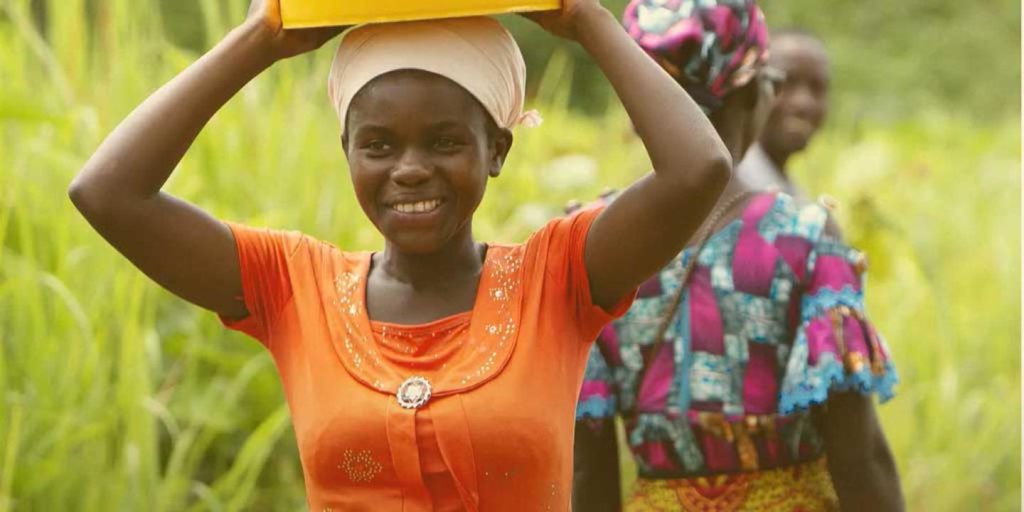 A woman carries a plastic bucket on her head and held by raised arms and hands.