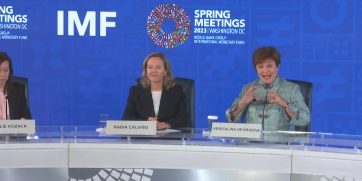 Two women sit behind a press conference desk against a backdrop, one listens as the other speaks and  gestures.