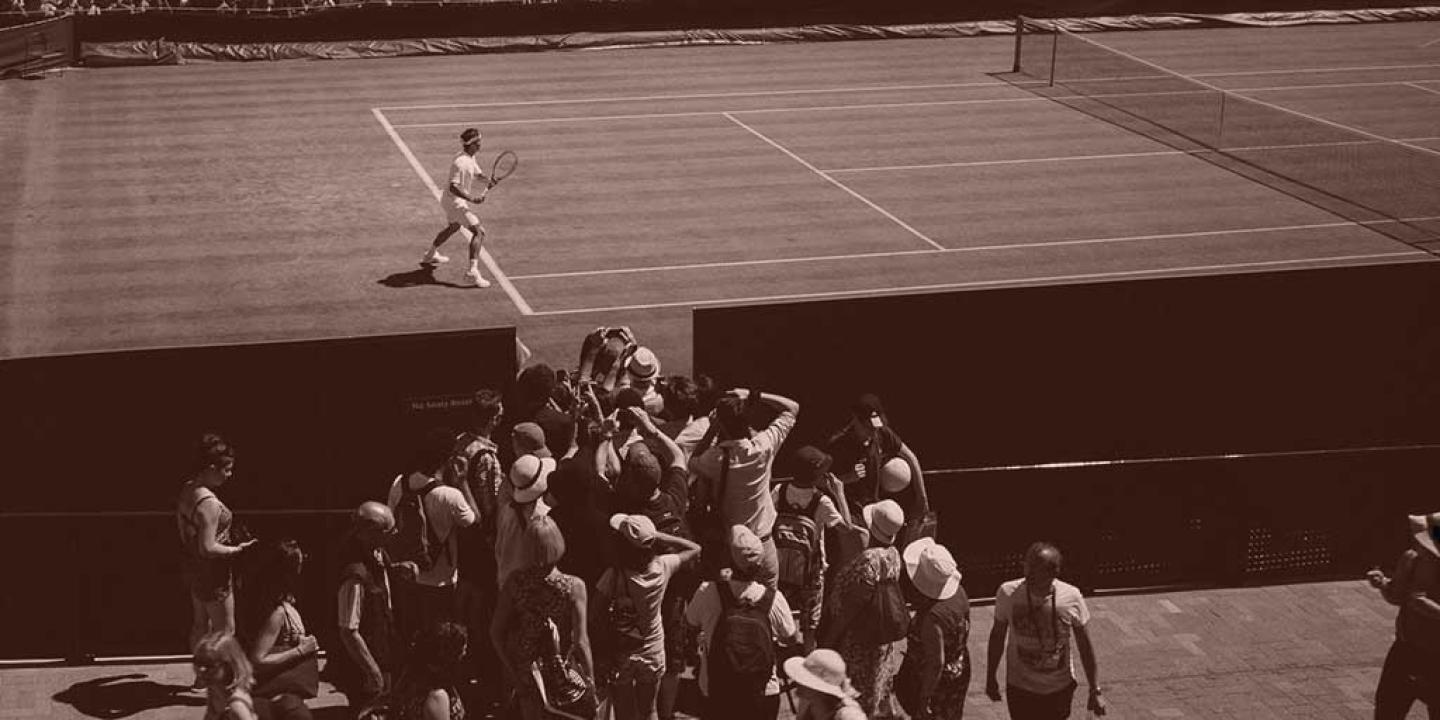 A tennis player stands ready to return a shot, while a phalanx of photographers crowd round a court-side opening to take a picture of him.