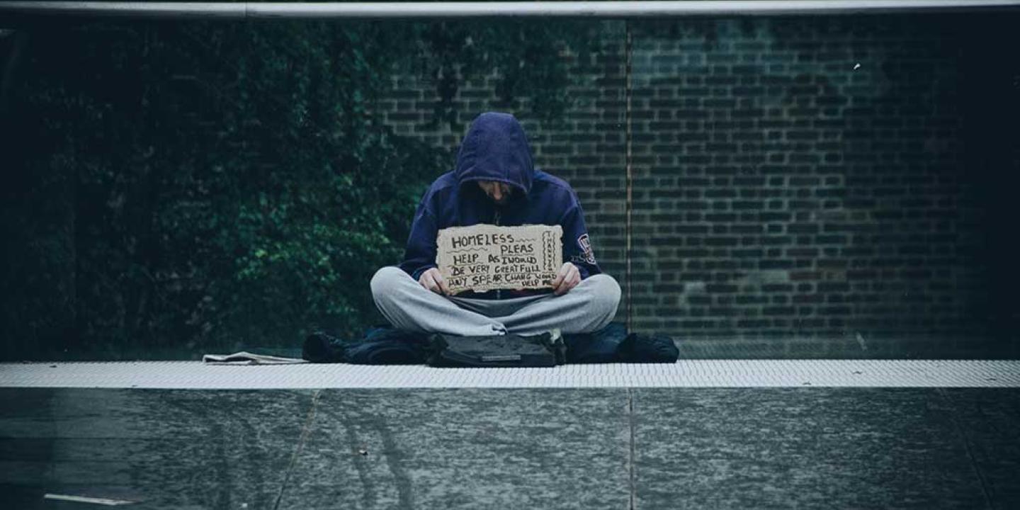 A beggar sits cross legged against a glass railing holding a sign.