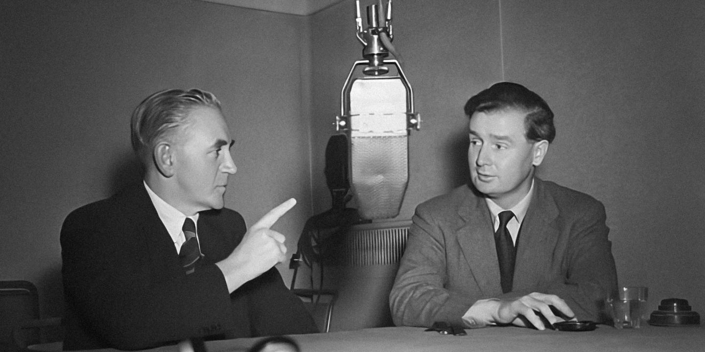 Two 1950's men un suits sit at a table dominated by a large hanging microphone. One points a raise hand and finger into the air. The other listens.