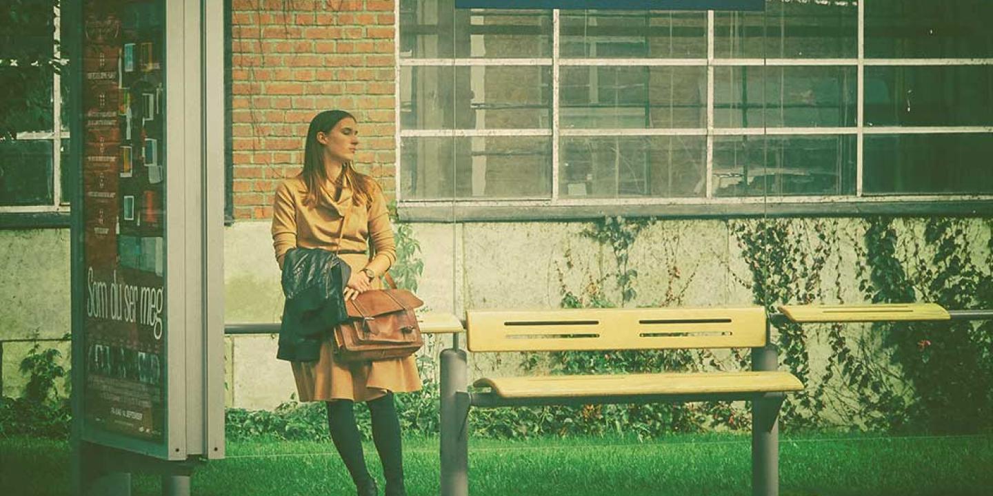 A woman leans against the glass of a bus shelter while waiting, she clasps a bag.