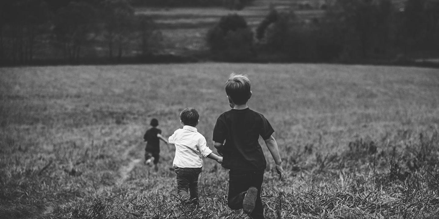 Three young children run away into the distance down across a field of long grass.