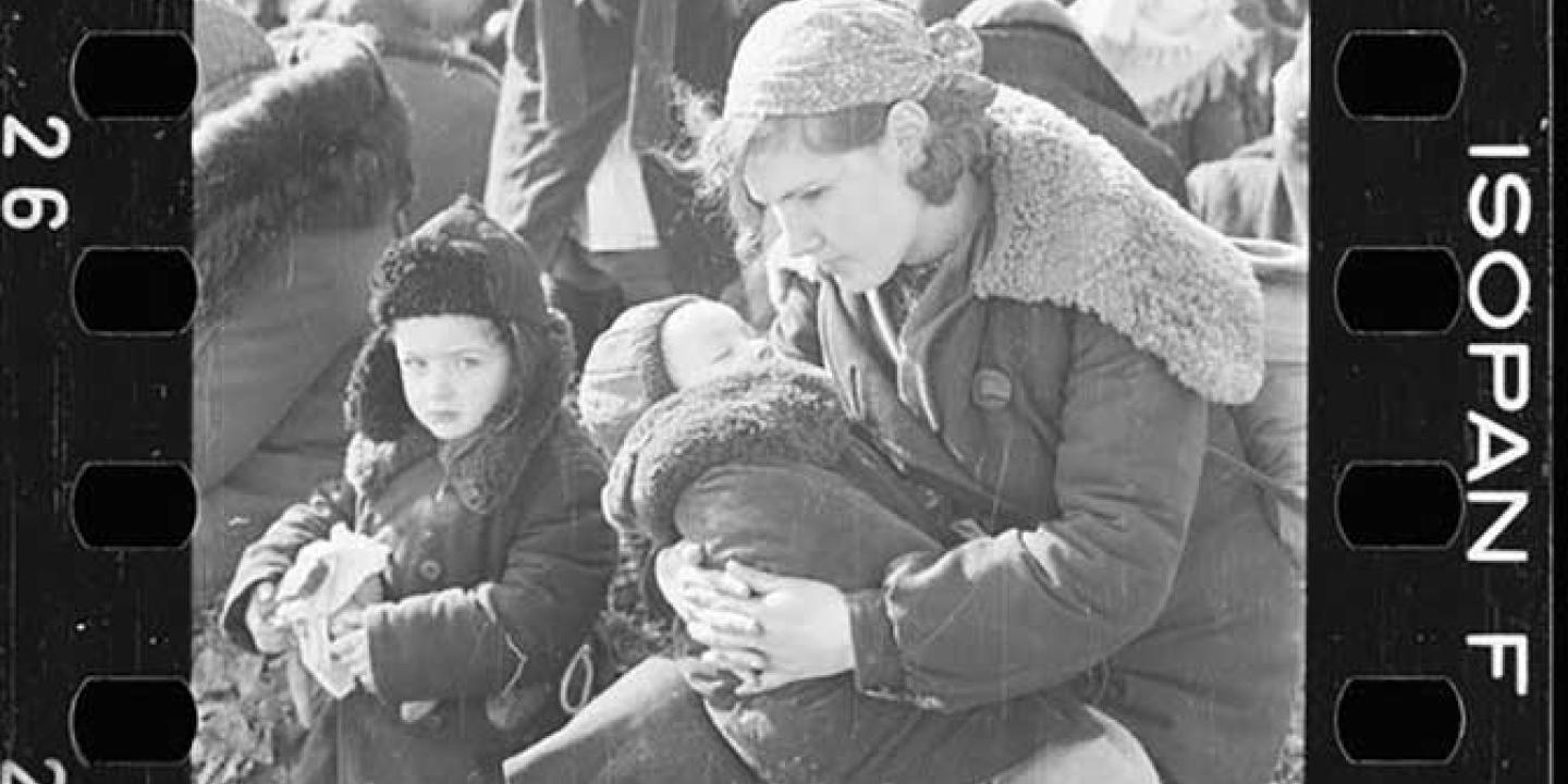 A mother cradles a child while another stands close by. They wear winter clothes of the 1940s and are amidst others waiting.