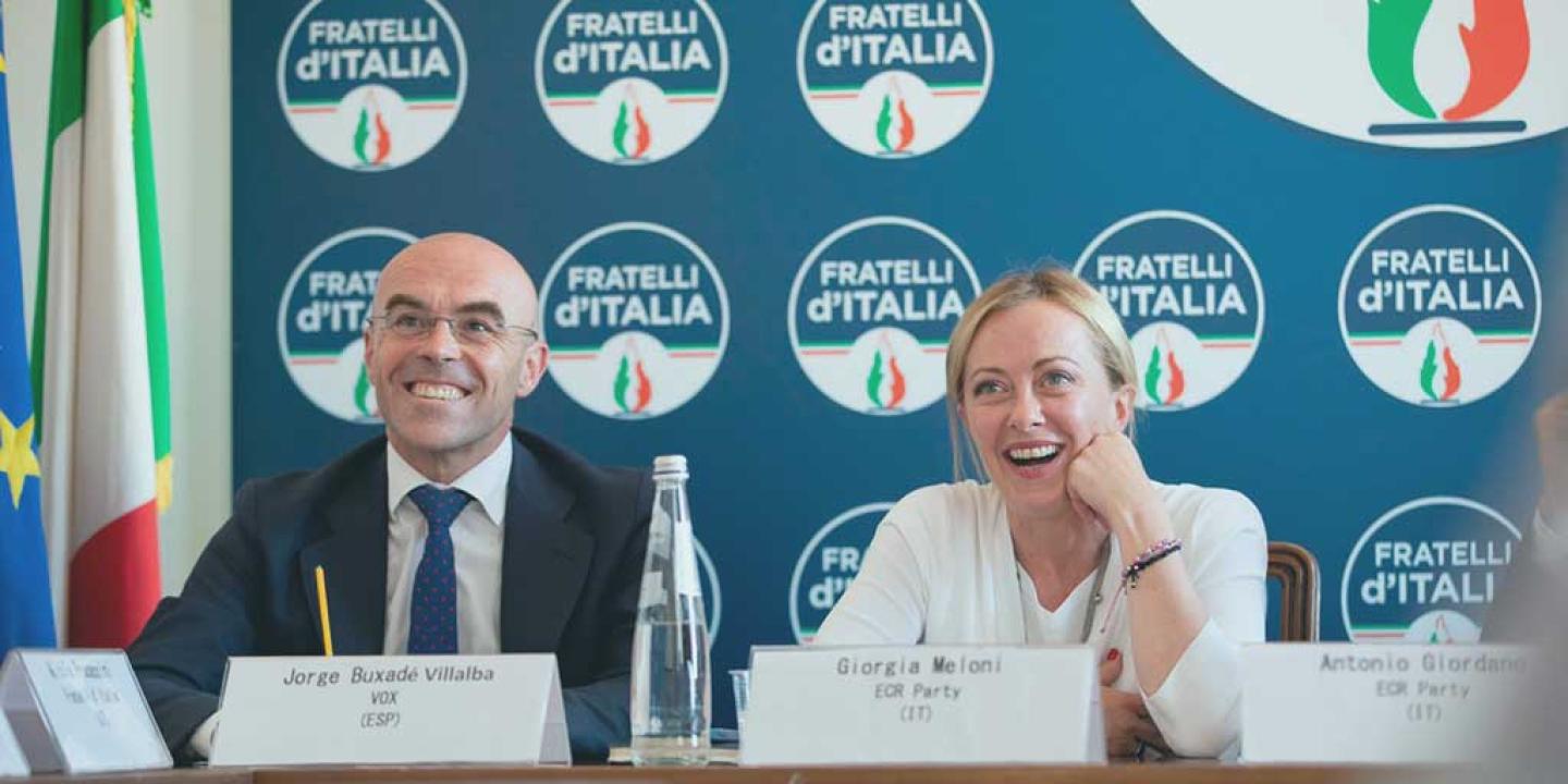 two politician site at a press conference desk and laugh, behind them is a backdrop of the political party's logo.