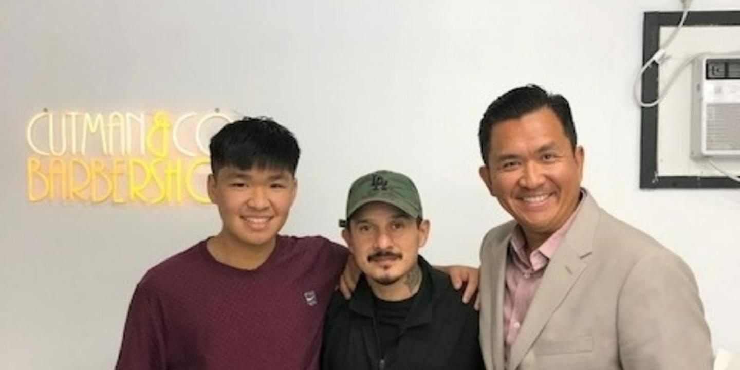 A barber stands between two clients, a father and son, a neon sign shines behind.