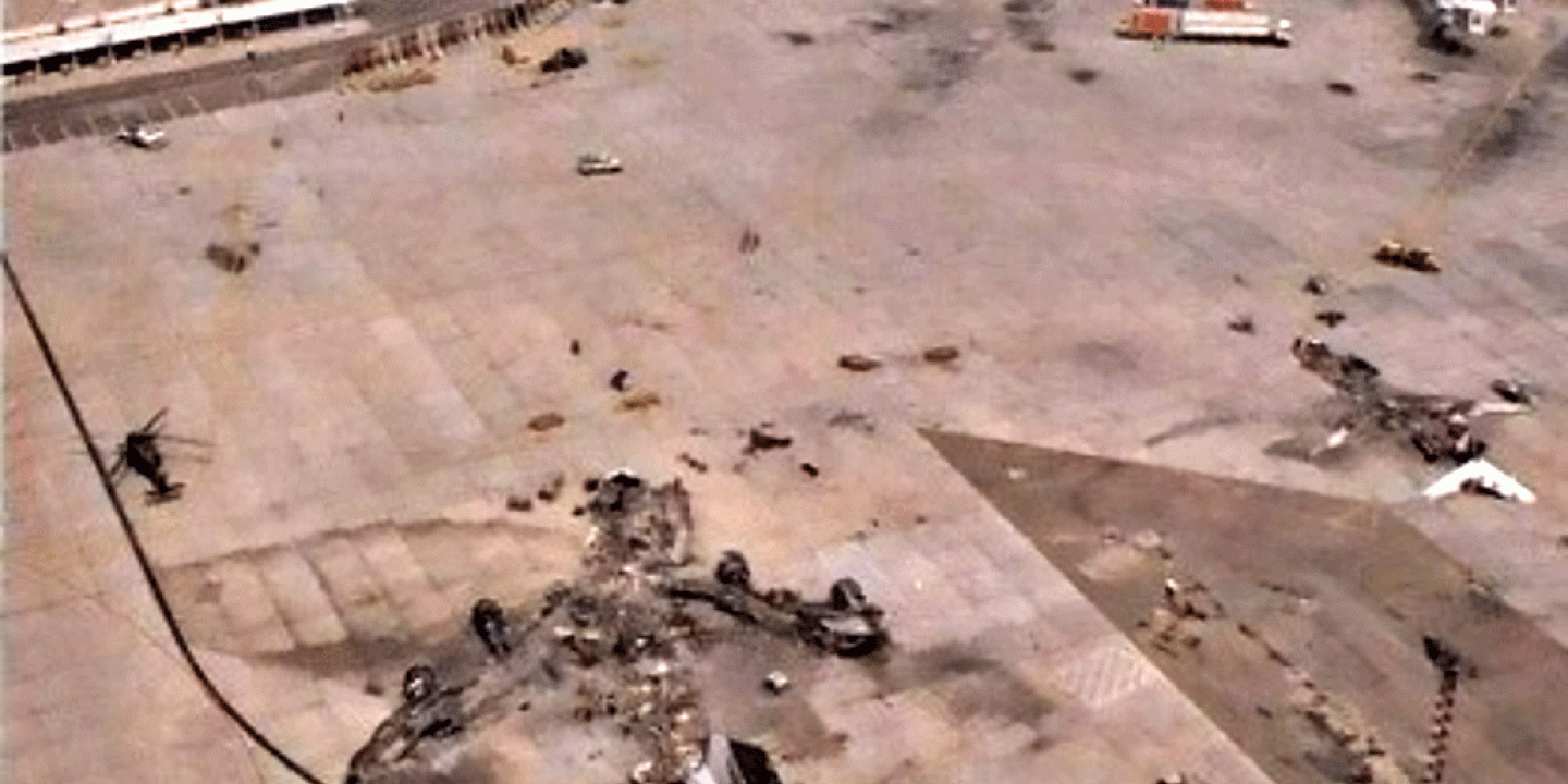 A destroyed airliner lies on the apron of a war-torn airport.