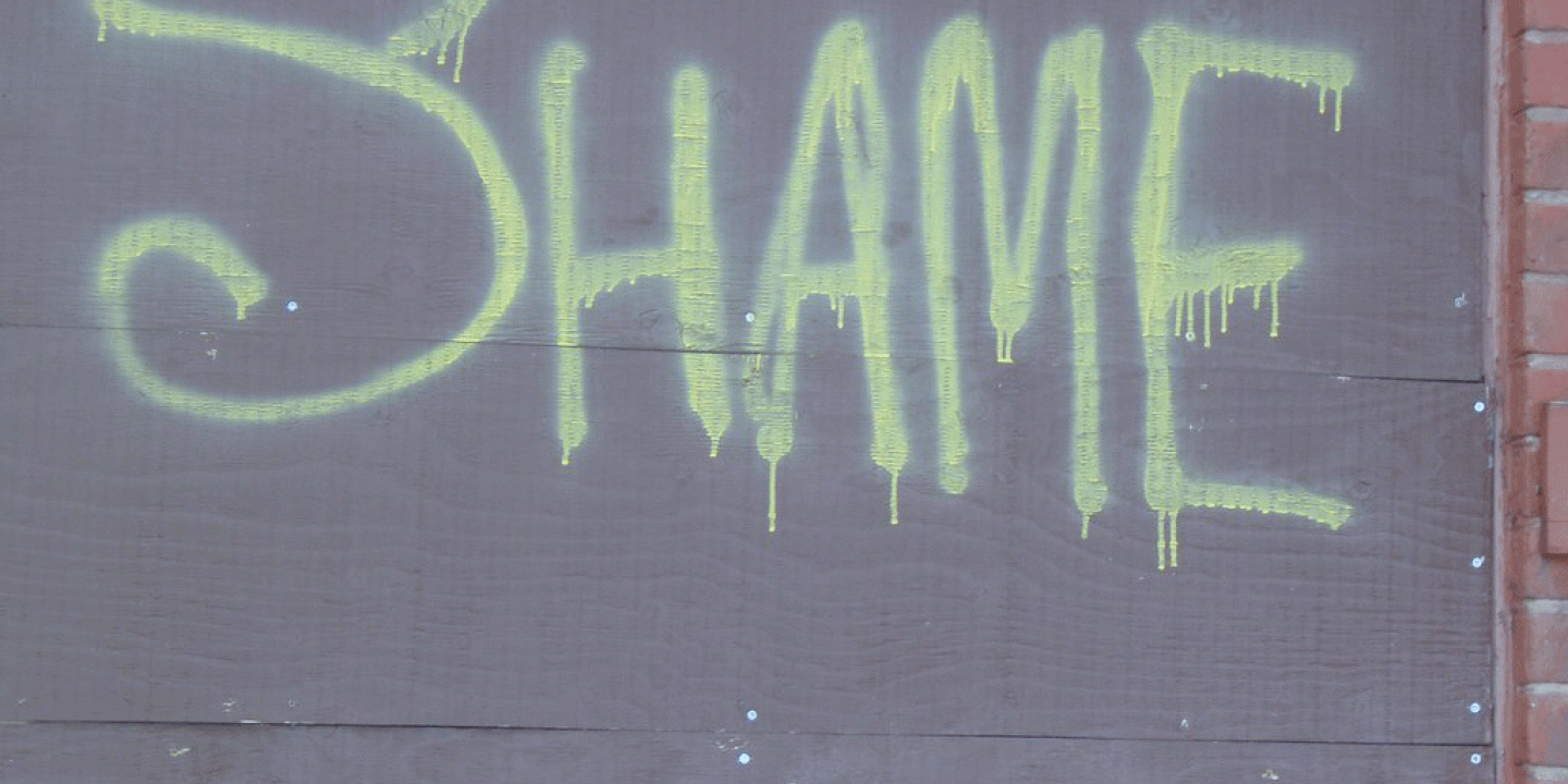 The word 'SHAME' spray painted onto a grey hoarding in lime green paint.