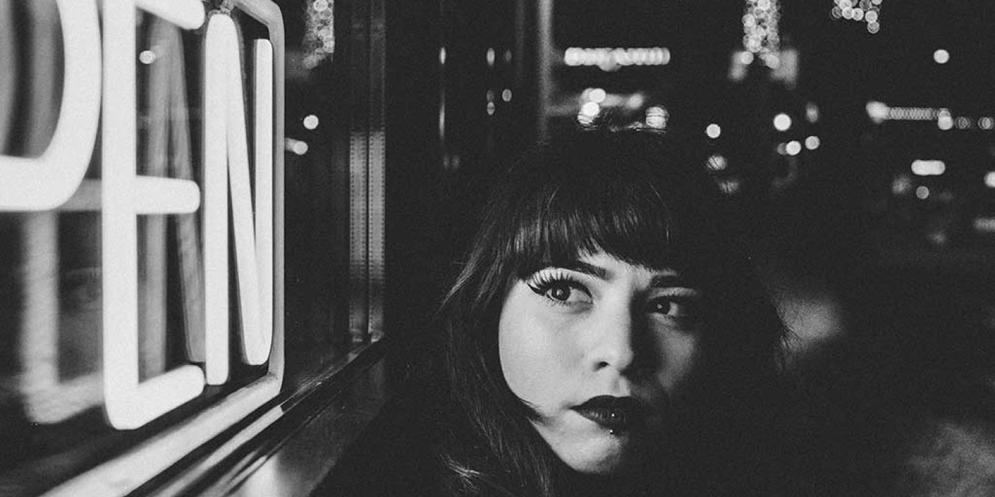 A black and white close up of a women in a street at night, turning to look around at a neion 'open' sign.