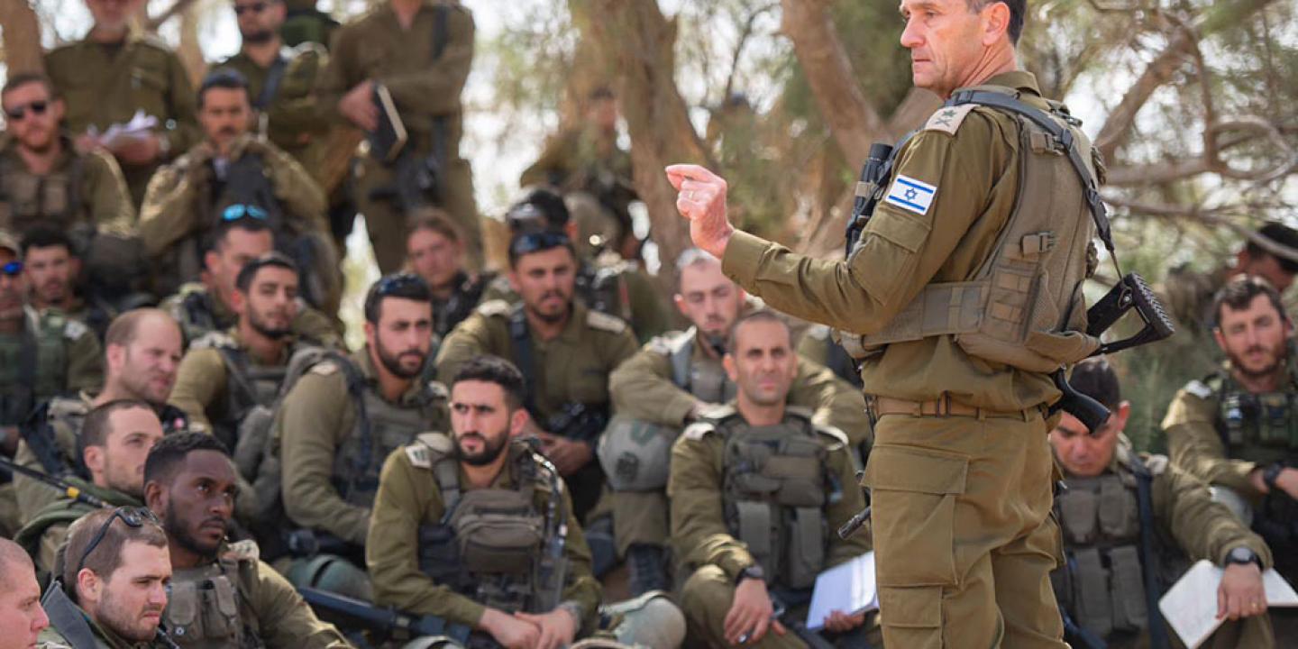 An army general in green fatigues stands and address a group of soldiers sitting, squatting and standing around him