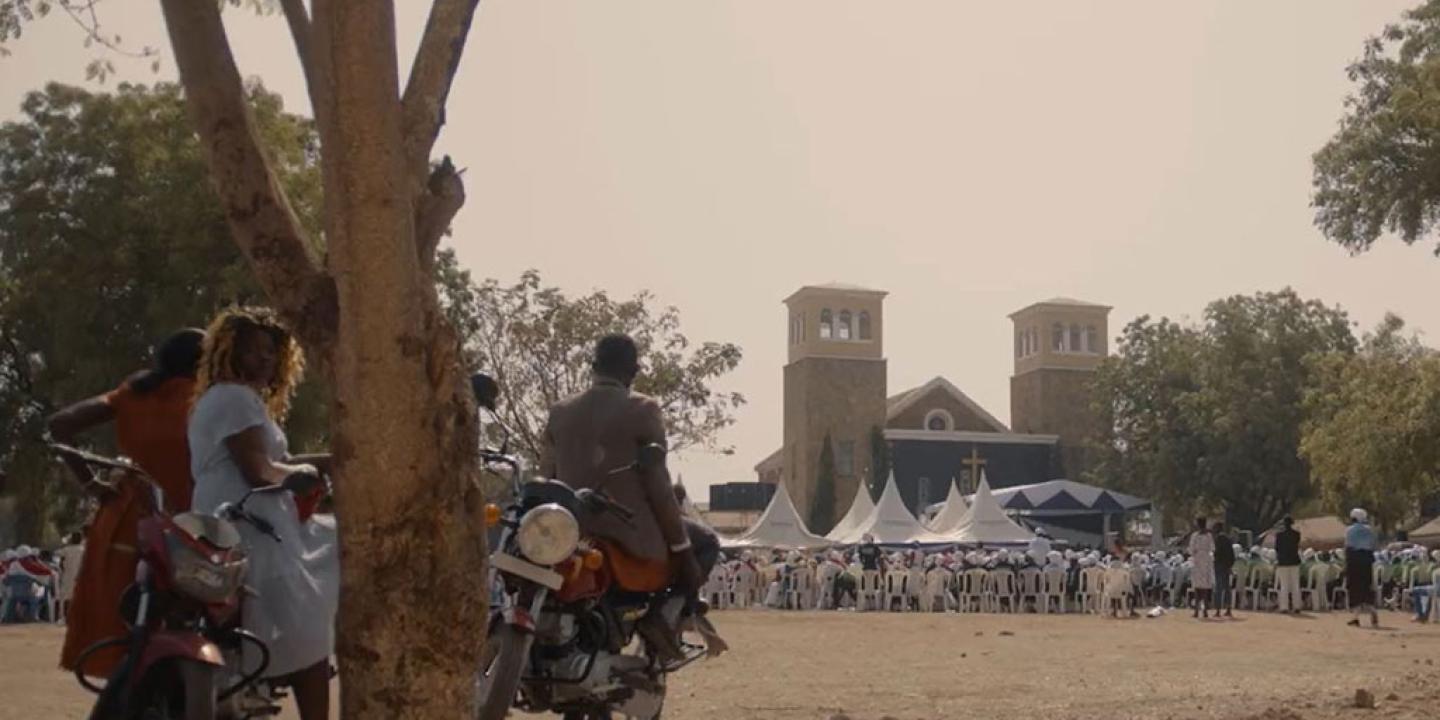 Outside a church a congregation waits seated while an onlooker rests on a motorcycle by a tree.