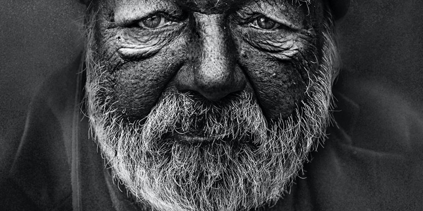 A black and white close up of the weather-beaten and wrinkled face and beard of a homeless man.