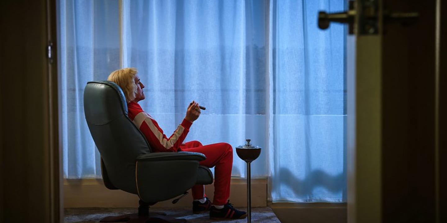 A man in a tracksuit sits in an arm chair smoking a cigar while looking towards a curtained window.