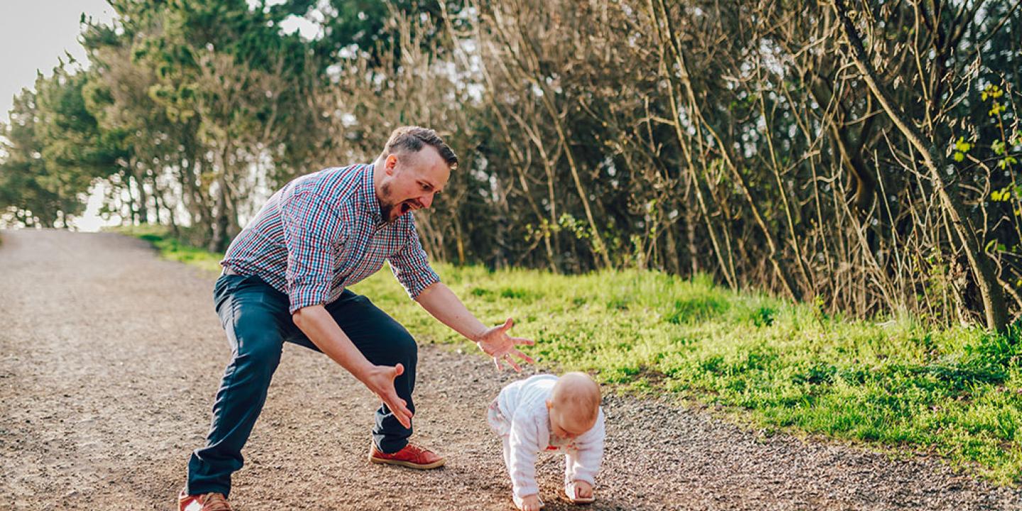 A dad hovers with open arms ready to catch a baby taking first steps