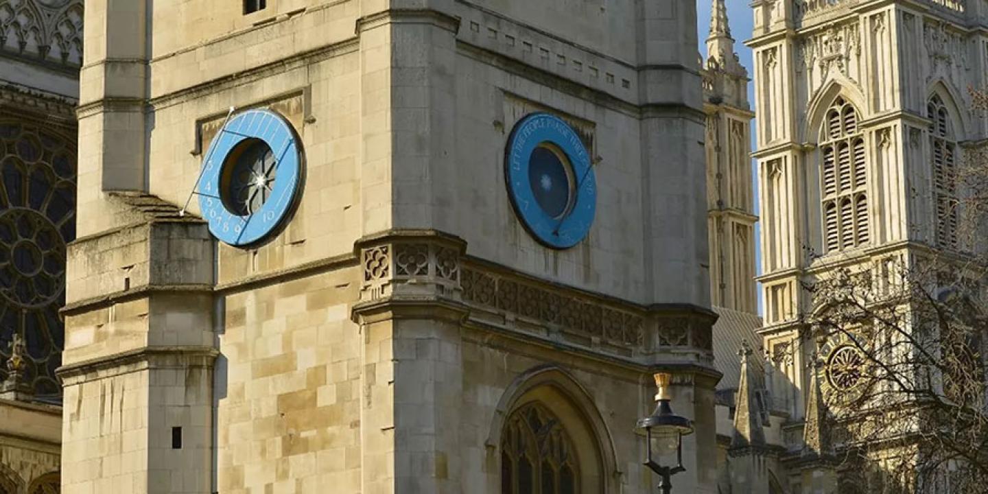 A close up of a blue sun dial on a church clock tower, with an abbey's windows behind it.