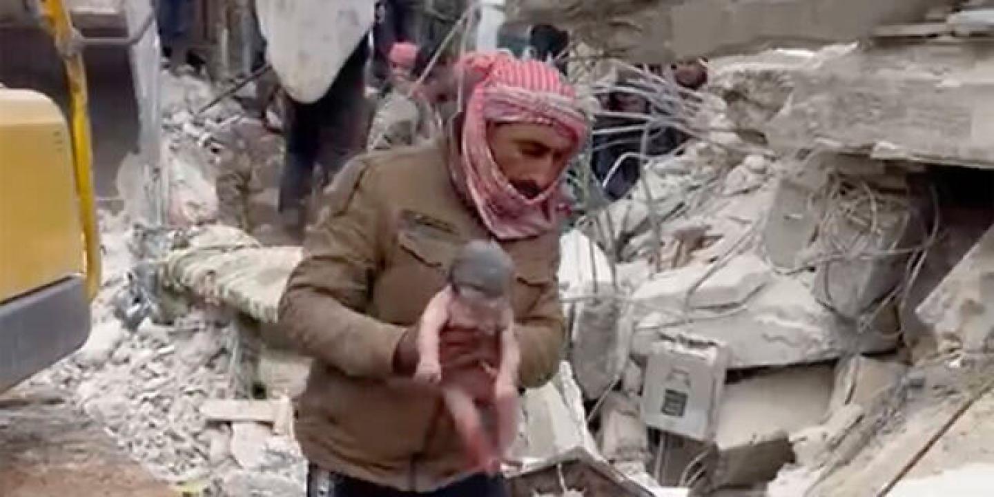 In front of a collapsed building, a rescuer carries a new born baby by the arms.