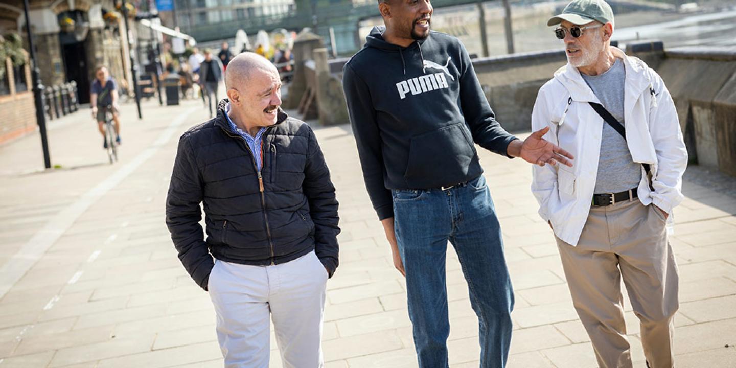 Three men walk down a path, the middle one talking and gesturing while the others listen.