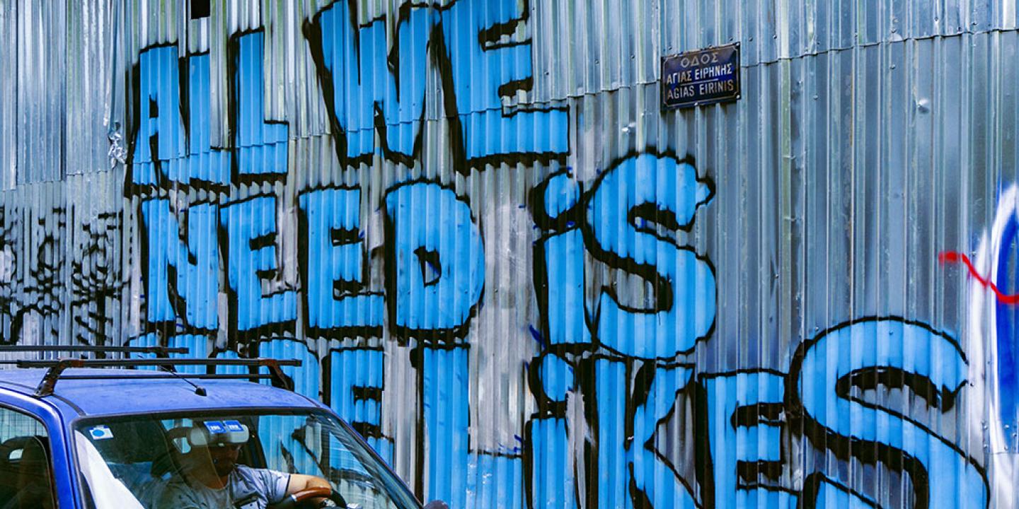 A corrugated sheet iron wall graffited in large blue letters that say 'All we need is more likes.'