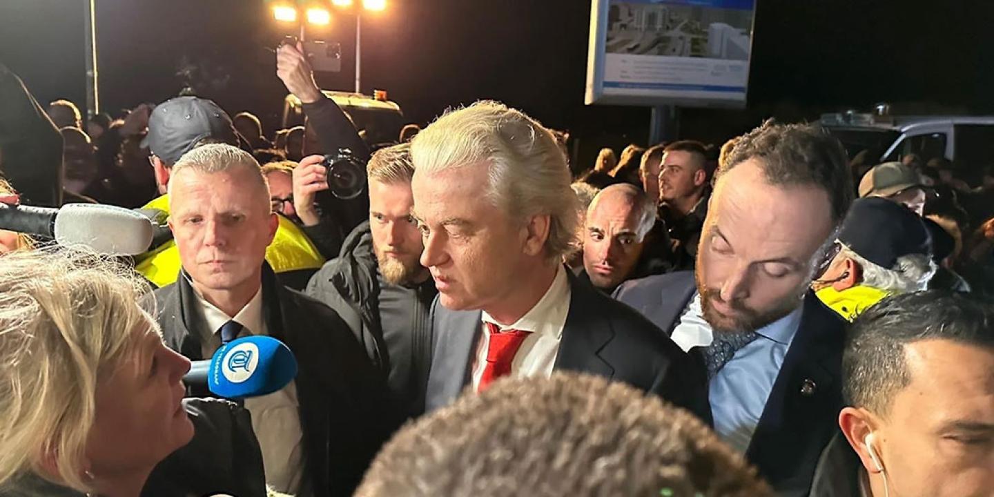 A politician in a suit stands amid a scrum of reporters holding microphones