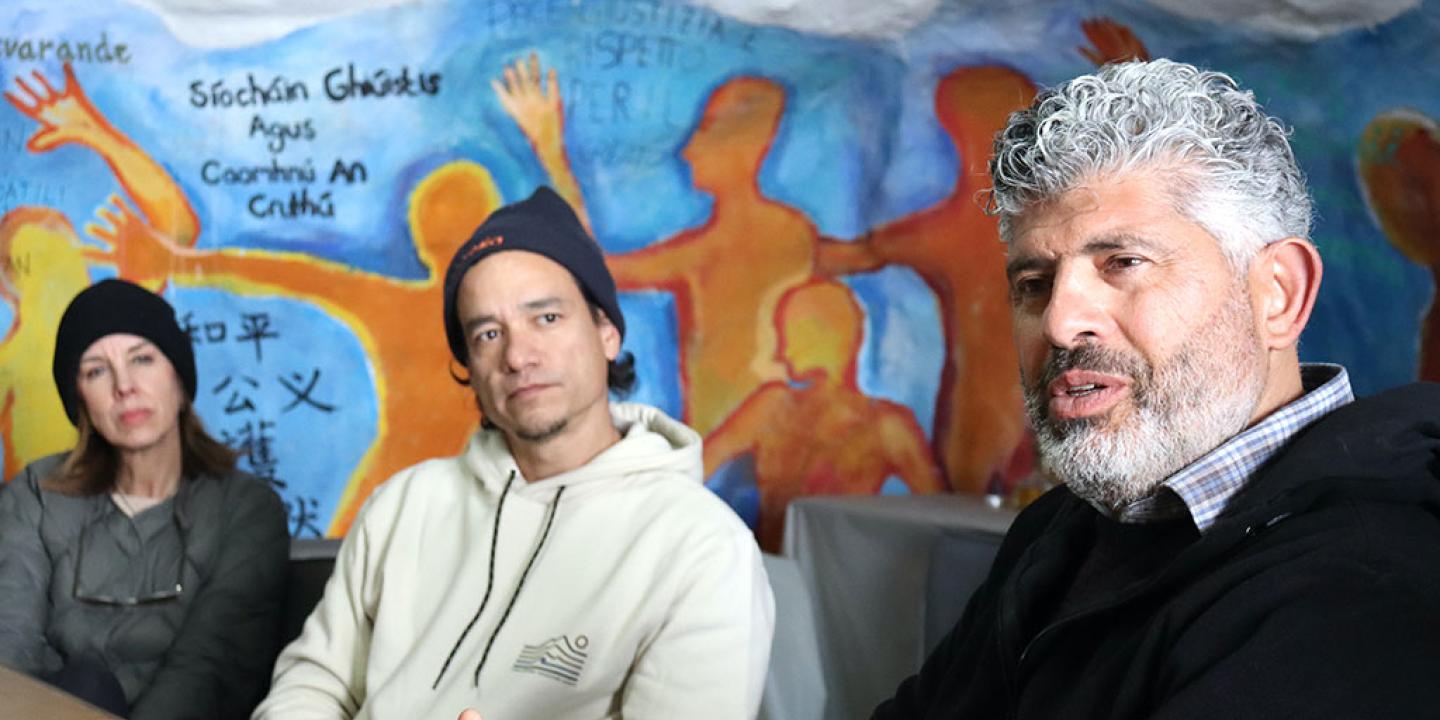 Two people down a table turn and listen to someone closer talk, against a wall mural.