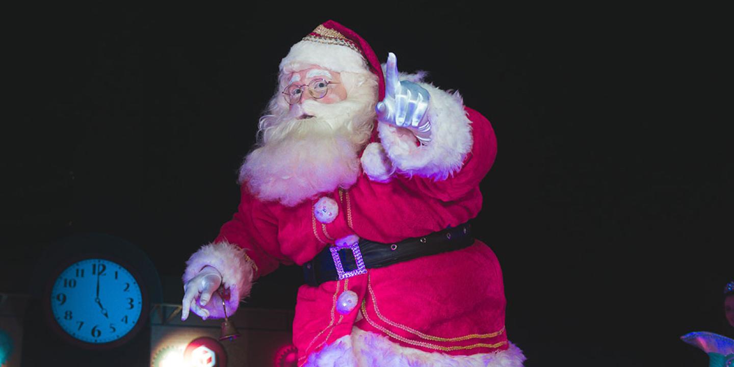 A Santa tiptoes in a darkened room carrying a lamp and holding up a finger in a gesture of silencing