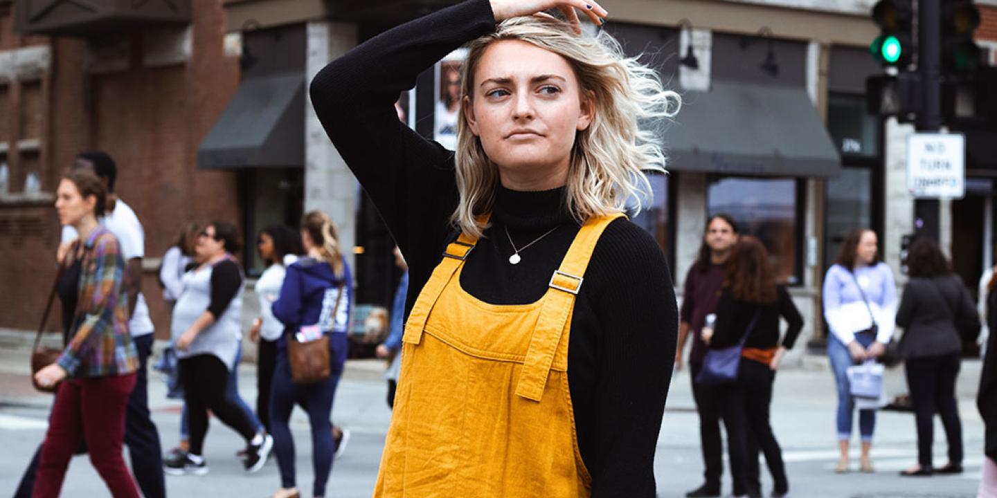 A woman stops in her stride down a street and pensively runs her hand through her hair as she looks to the side.