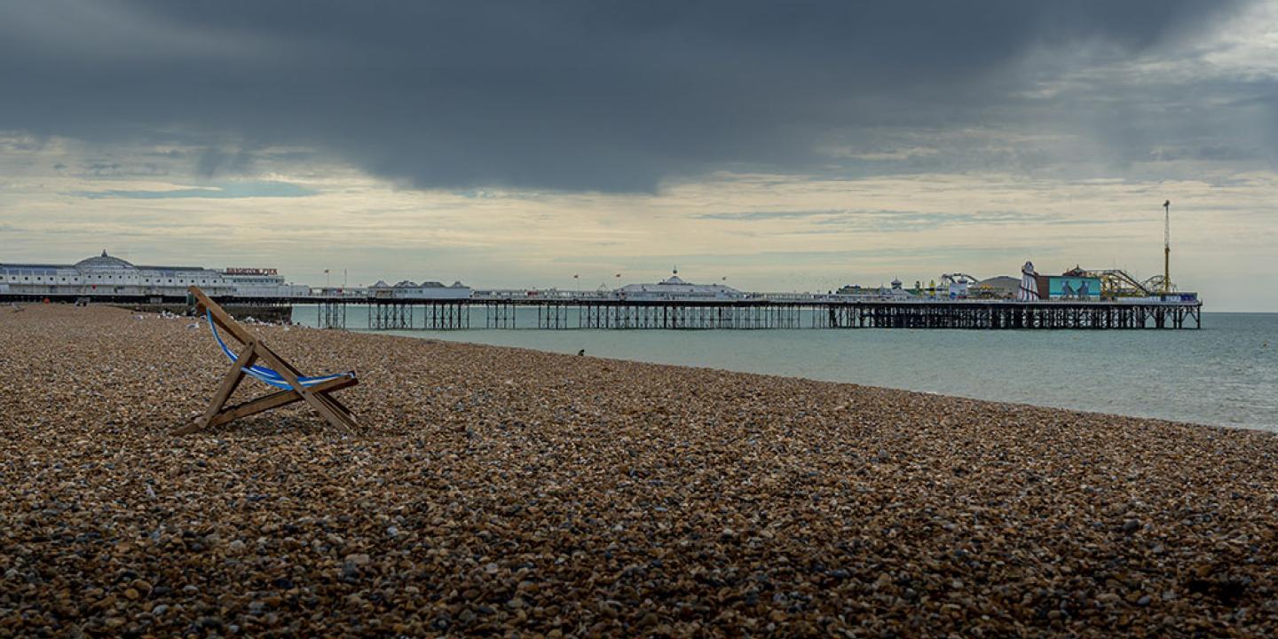 A moody sky overshadows a shingle beach on which a lone empty deckchair stands. A pier with funfair is in the middle distance.