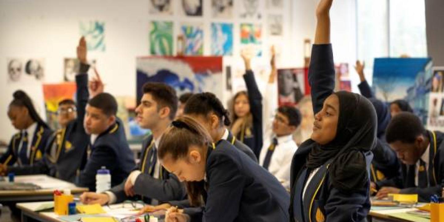 school pupils sit at desk, some with a hand raised.