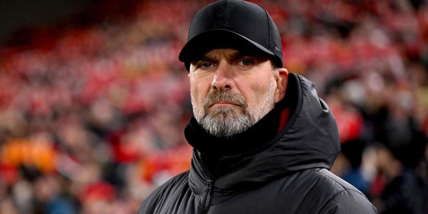 A football manager stands on the touchline and stares hard, dressed in a black coat and hat.