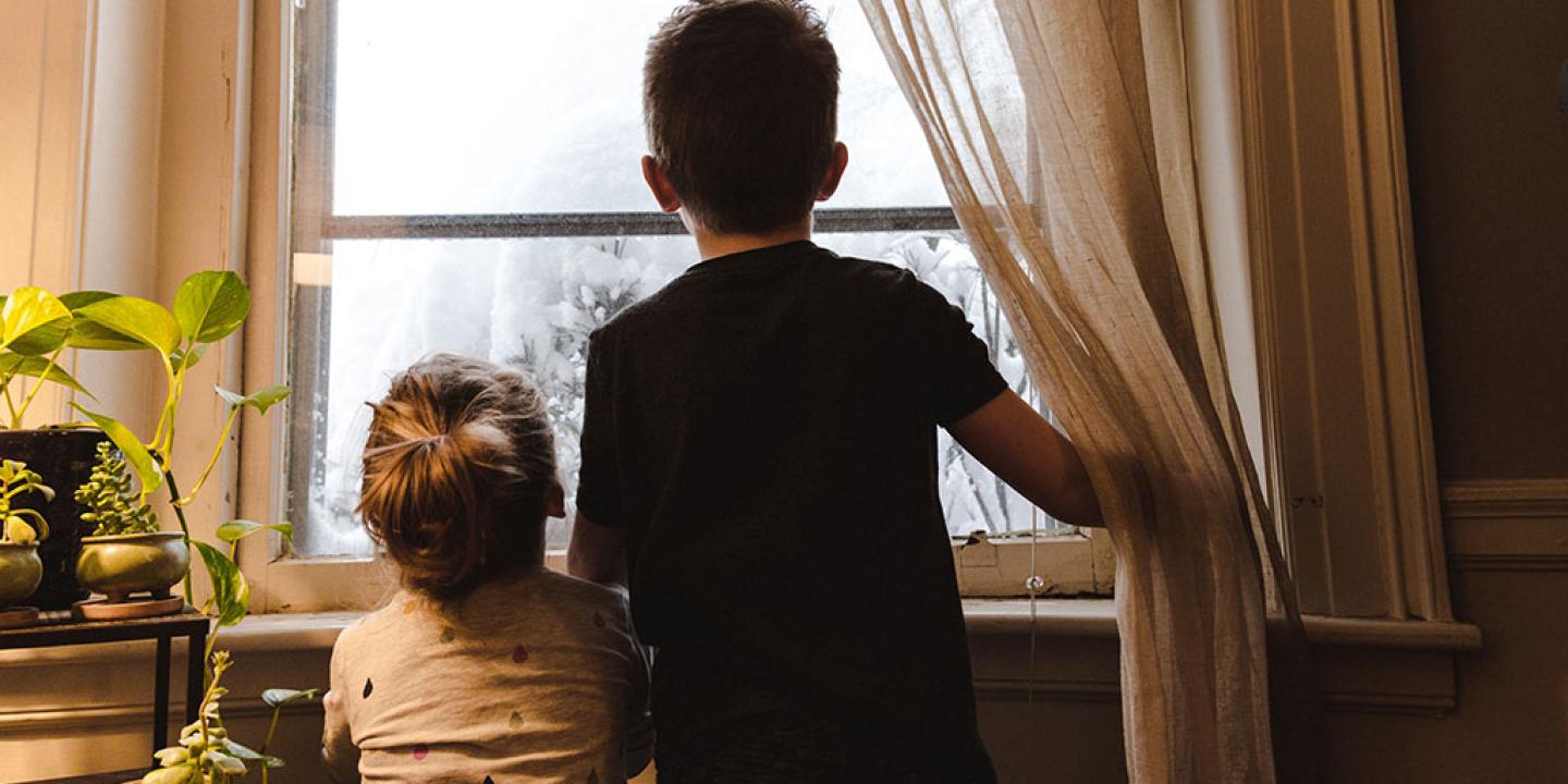 Two small chidlren push a curtain aside to stare out the window at snow.