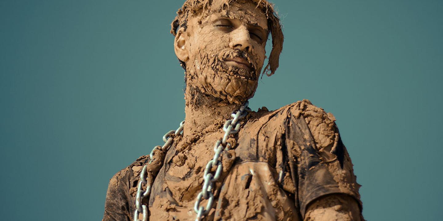 A man covered in dried and caked mud stands and looks to the side, a steel chain is draped from his shoulders.