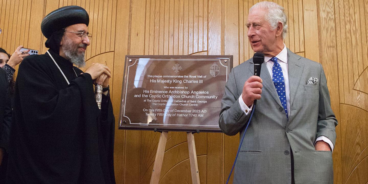 An archbishop wearing a black hat and robes stands next to a new building's plaque, while King Charles, wearing a suit stands the other side holding a mic.