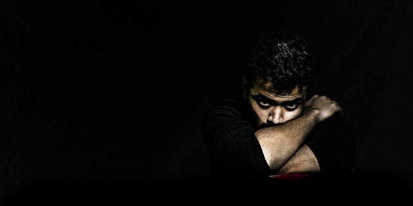 In a darkened room, a man's angry face is lit as he rests on arms folded tightly around it.