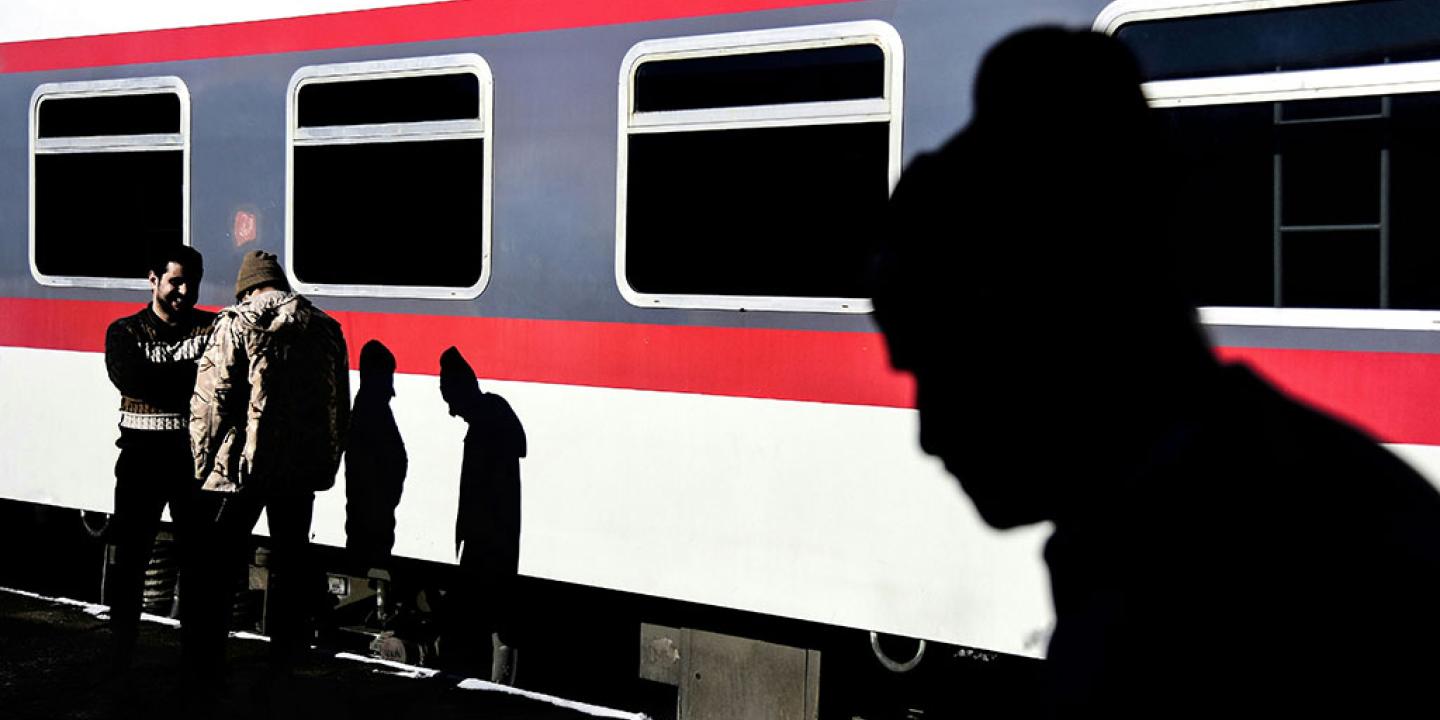 On a sun-lit railway platform, two men talk casting shadows on a waiting red and white train. Another shadow beside them is that of a man silouheted in the foreground